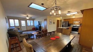 Uma sala de estar ampla com uma grande mesa de jantar de madeira, um sofá e uma área de cozinha com armários e eletrodomésticos de madeira.