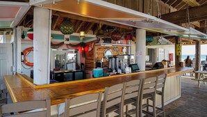 Un bar en la playa con mobiliario de madera, una caja registradora y diversos utensilios de bar.