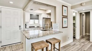 Una cocina moderna con encimeras de granito, horno empotrado y pileta de acero inoxidable. Hay dos taburetes de madera y un cuadro en la pared.