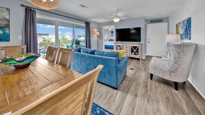A modern living room with a wooden dining table, blue sofa, and a TV. - Blue Haven- Beachside condo - 4 minute walk to sand! (Santa Rosa Beach)