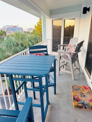 Un balcon abrité avec une table et des chaises bleues, un tapis coloré et une vue sur des maisons et des arbres.