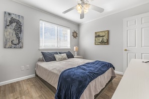 Quarto com cama, ventilador de teto, janela com persianas, relógio de parede e porta.