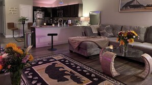 Ein modernes Wohnzimmer mit einer Couch, einem Couchtisch und einem Teppich. Im Hintergrund ist eine Küche mit dunklen Schränken und einem Kühlschrank zu sehen.