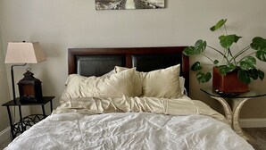 Ein ordentlich bezogenes Bett mit weißer Decke und Kissen, eine Nachttischlampe, ein gerahmtes Bild an der Wand und ein Blumentopf auf einem Beistelltisch.