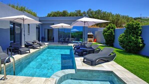 A modern outdoor pool area with lounge chairs and umbrellas, surrounded by a well-maintained lawn and a barbecue area.