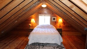 A cozy attic bedroom with a wooden sloped ceiling, a bed with a white comforter, two bedside lamps, and a window with natural light. - Rustic Chapparal Cabin at Bass Lake Heights (Oakhurst)