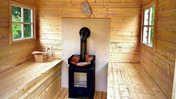 A wooden cabin interior with a wood-burning stove, benches, and windows. - Rosenlund - Holiday cottage Rosenlund (Älmhult)