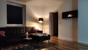 A modern living room with a dark-colored sofa, a small round table, a lit candle, and a floor lamp. A flat-screen TV is mounted on the wall.