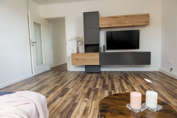 A modern living room with a flat-screen TV mounted on a wall, wooden and dark grey storage units, a round wooden coffee table with candles, and a wooden floor. - Beautiful vacation apartment in Oberkotzau am Fichtelgebirge (Oberkotzau)