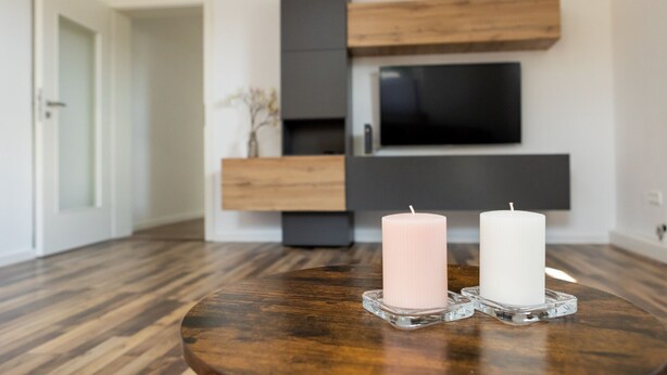 A modern living room with a wooden floor, a TV mounted on a wall, and two candles on a glass table.