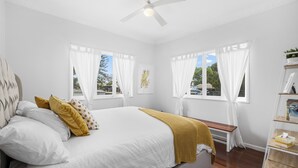 Une chambre à coucher avec un lit, des rideaux blancs, un ventilateur de plafond et une fenêtre donnant sur l’extérieur.