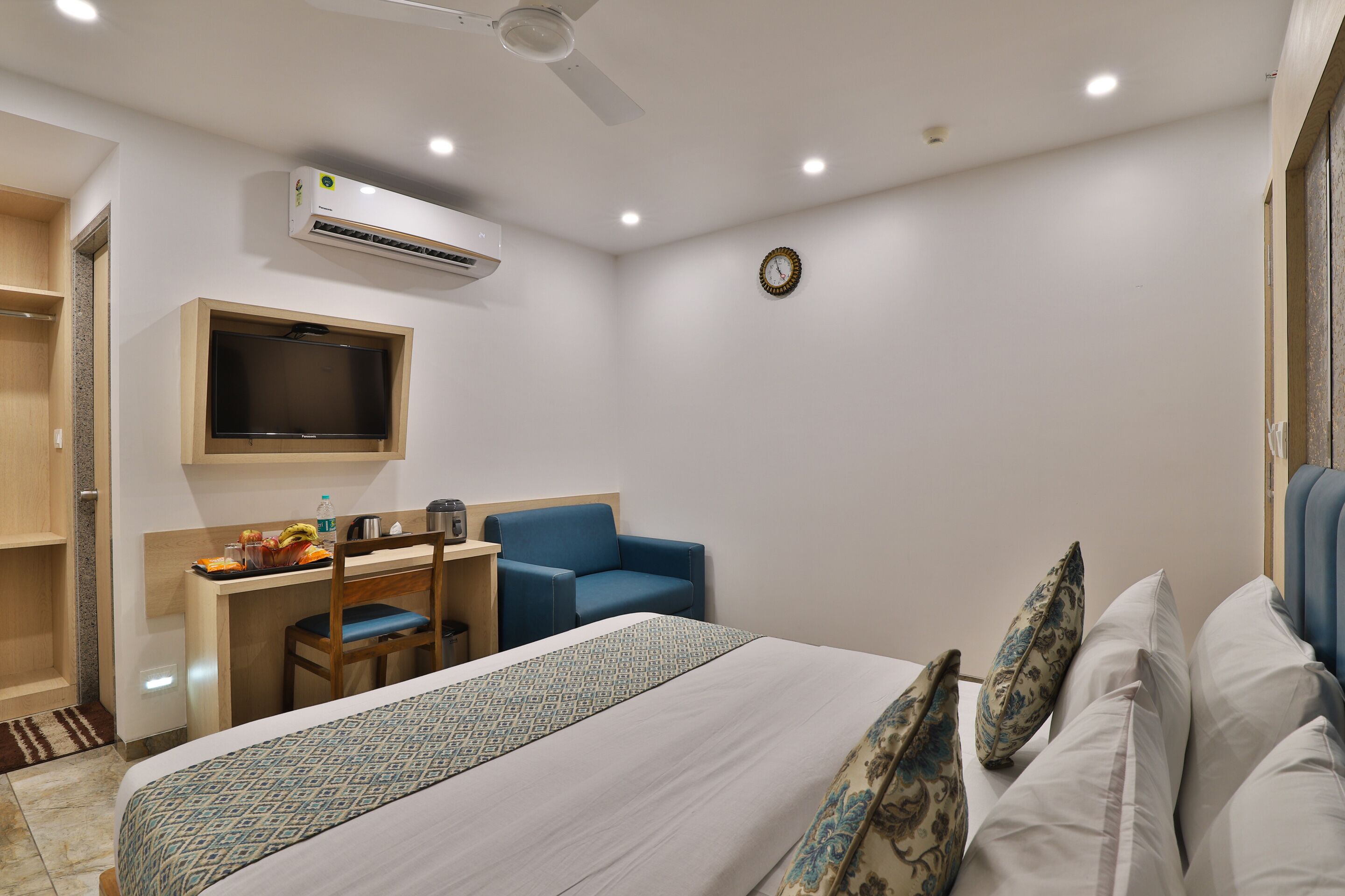 Habitación de hotel con cama, escritorio con silla, televisión, equipo de aire acondicionado y un reloj en la pared.