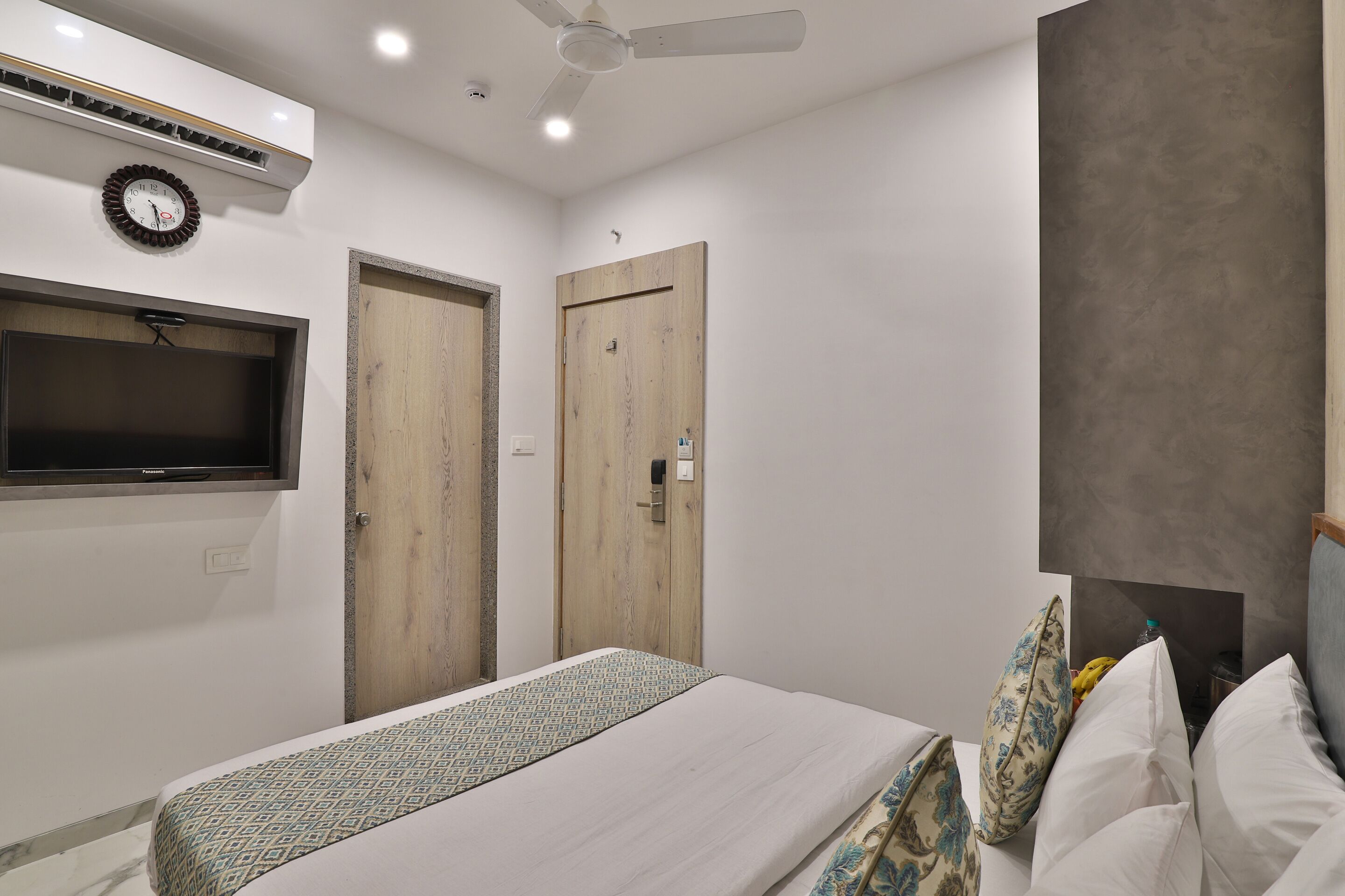 A bedroom with a bed, a TV mounted on the wall, two wooden doors, and a ceiling fan.