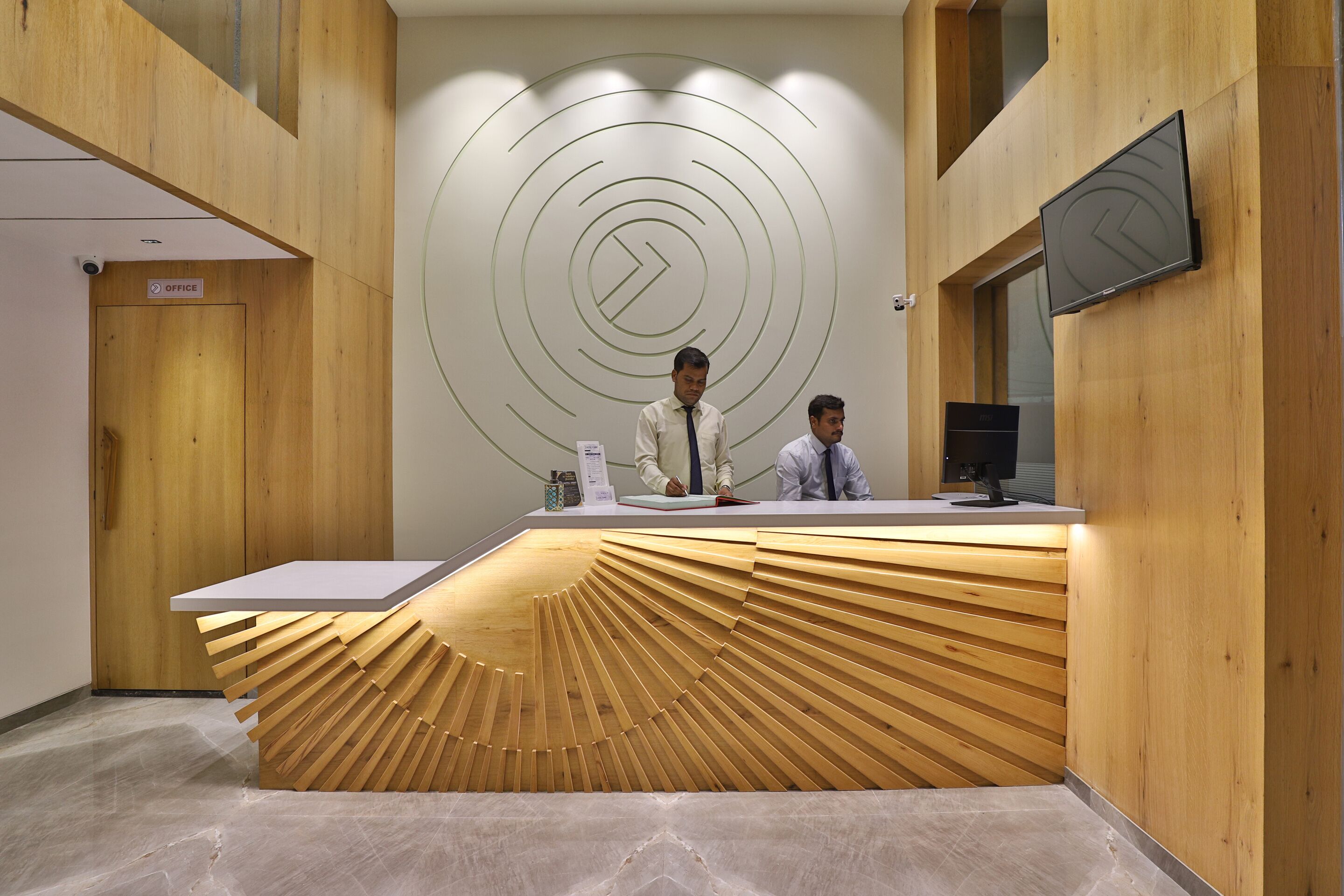 A reception desk with a unique wooden design, two staff members, and a modern wall with circular patterns.