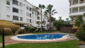 A swimming pool surrounded by a well-maintained lawn, with a building featuring balconies and a thatched roof structure in the background. - Benalmar Playa (Benalmádena)