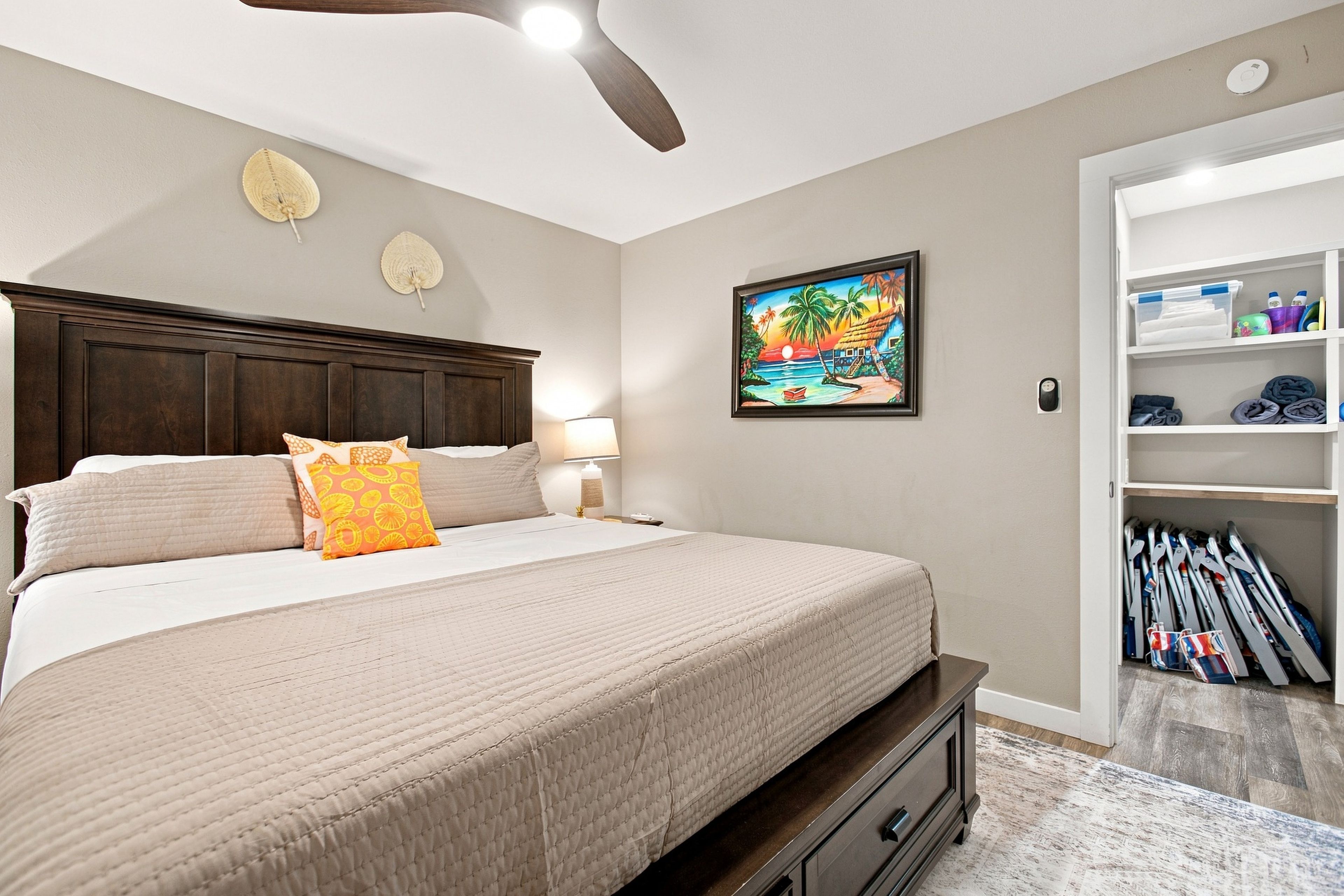 A bedroom with a bed, a nightstand, a lamp, a ceiling fan, a painting, and a closet with folded clothes.