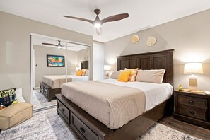 A bedroom with a large bed, a ceiling fan, a nightstand, a lamp, a mirror, and a painting on the wall.