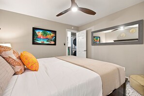 A bedroom with a bed, a ceiling fan, a painting of a tropical scene, a door leading to a laundry area, and a mirror on the wall.