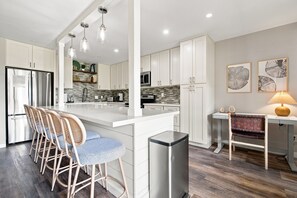 A modern kitchen with white cabinets, a central island with bar stools, a microwave, and a coffee machine.