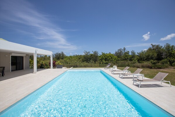 A rectangular swimming pool with clear blue water, surrounded by a white deck. There are lounge chairs aligned along the pool's edge.
