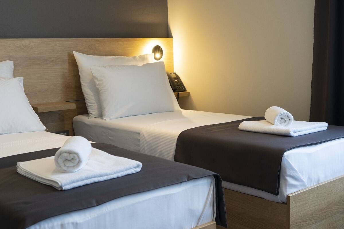 Ein Hotelzimmer mit zwei Betten, jeweils mit weißen Bettwäsche und einer dunkel farbigen Tagesdecke. Ein Nachttisch mit Lampe und Telefon steht daneben.