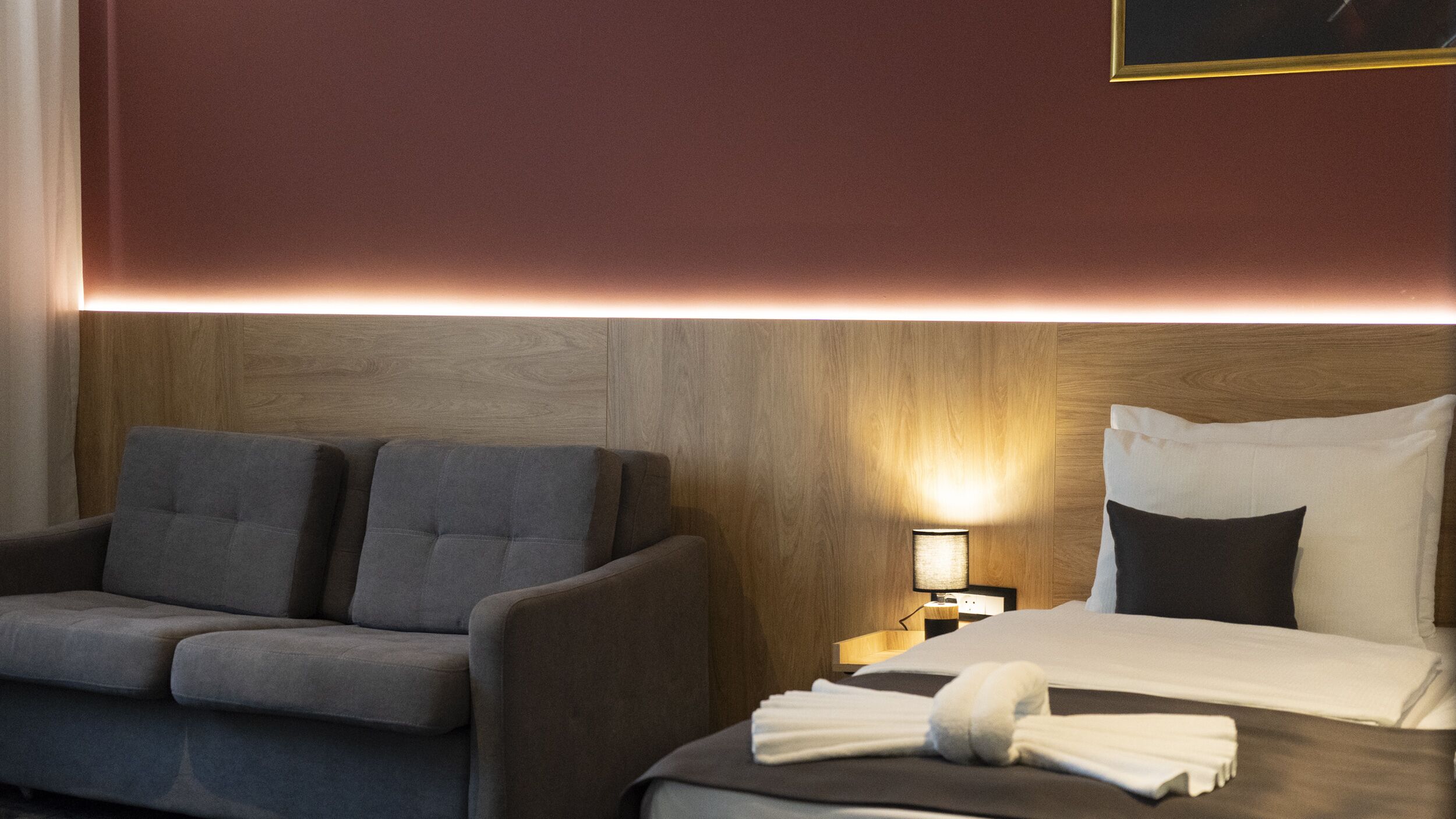 A hotel room with a sofa, a bed with a neatly made bed and a towel, a nightstand with a lamp, and a wooden headboard.