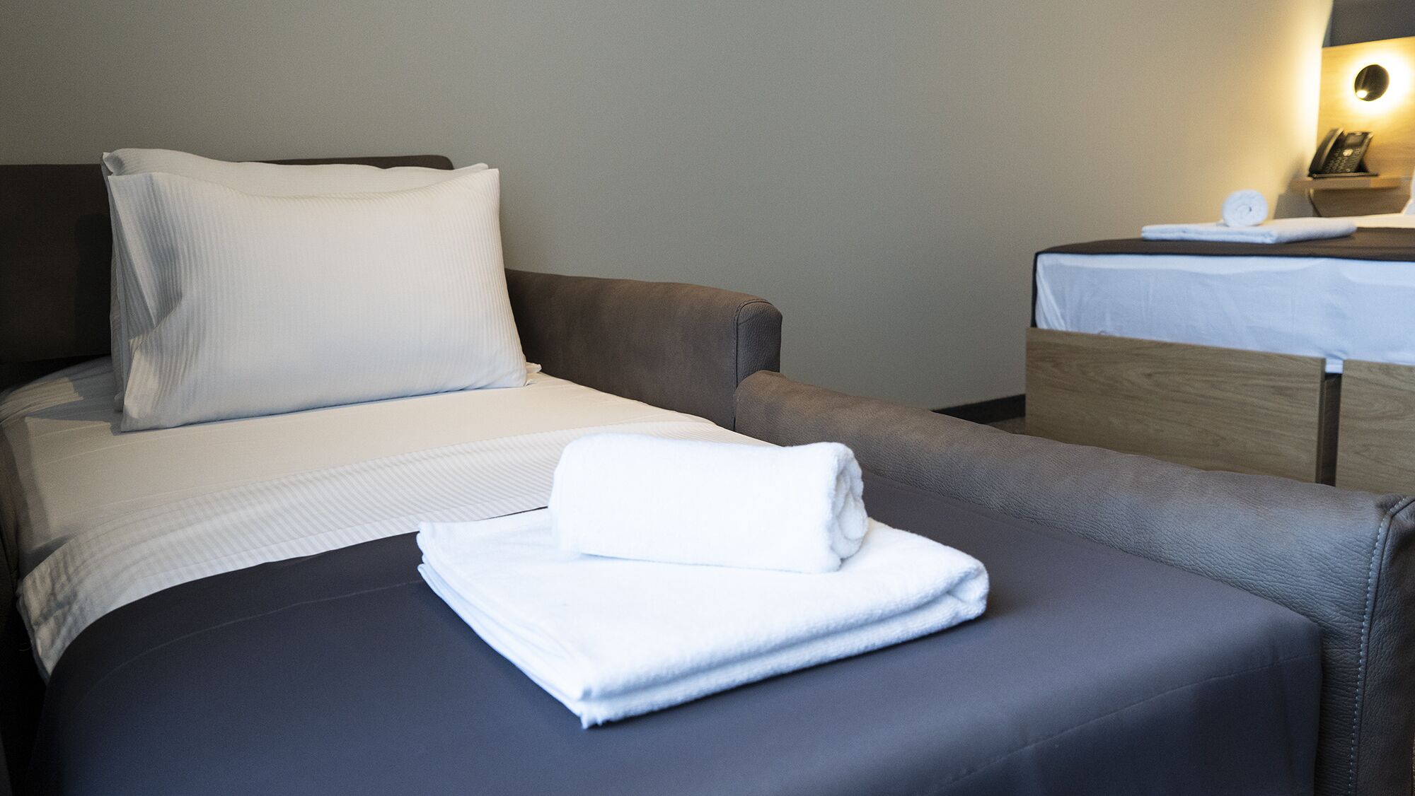 A hotel room with a bed, a bedside table with a lamp, and a folded towel on the bed.