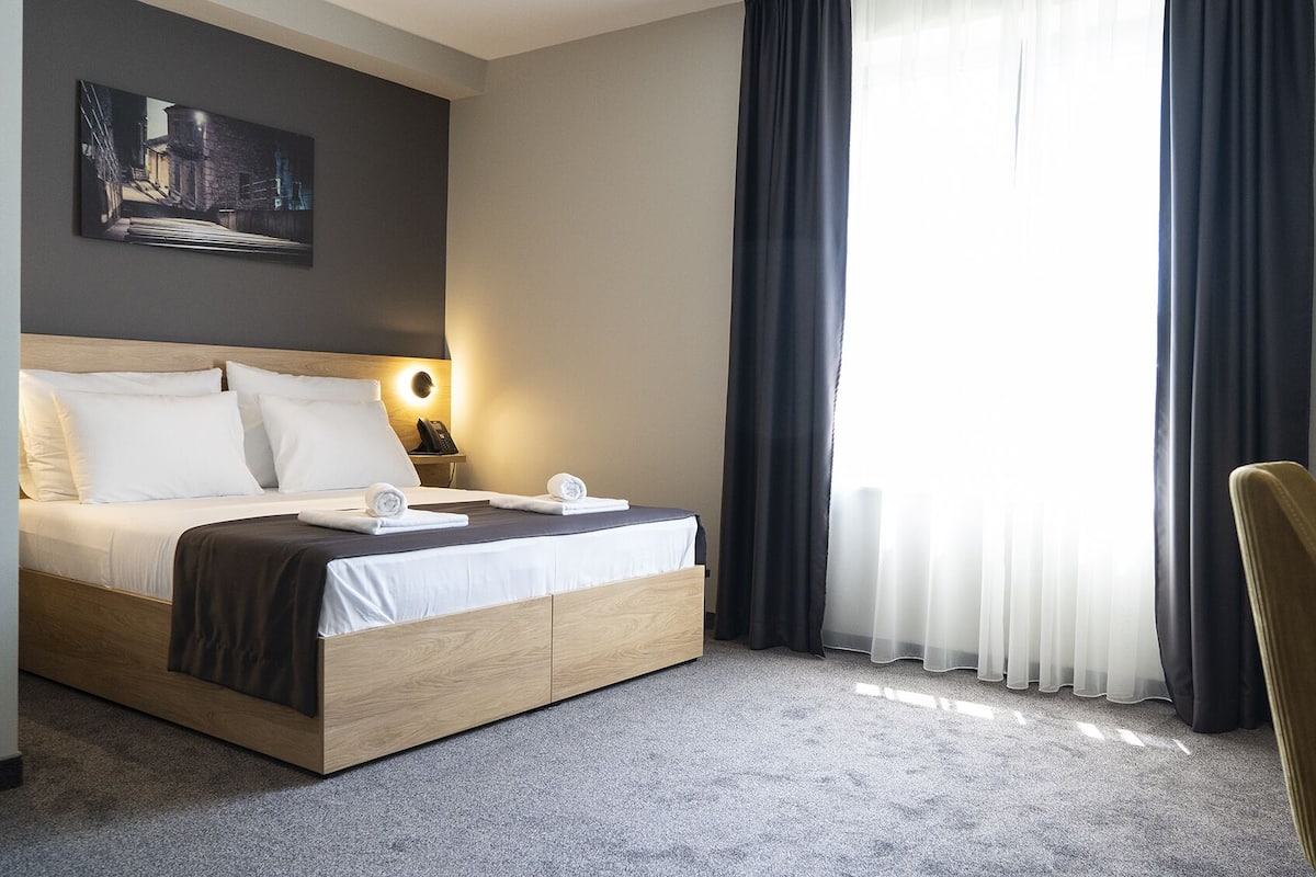 Ein Hotelzimmer mit Bett, Schreibtisch, Stuhl, einem Bild an der Wand und einem Fenster mit Vorhängen.