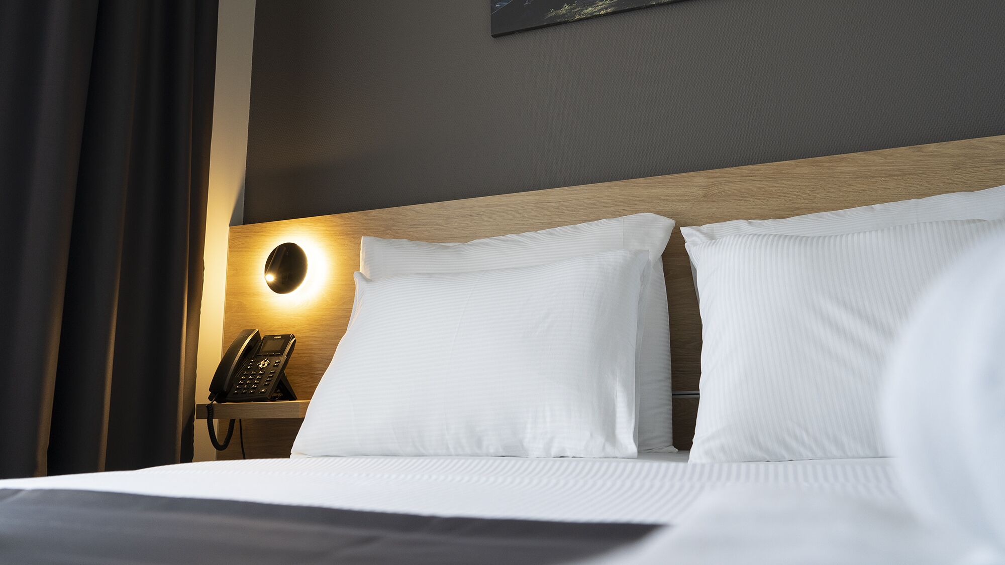 A hotel room with a neatly made bed, a bedside telephone, and a wall-mounted light fixture.