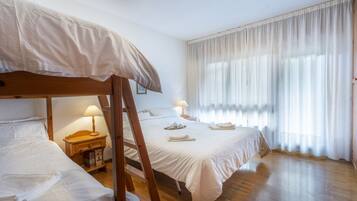 Una camera d'albergo con un letto a castello, un comodino con una lampada, una finestra con tende leggere e un pavimento in legno.
