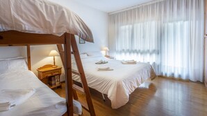 A hotel room with a bunk bed, a bedside table with a lamp, a window with sheer curtains, and a wooden floor.