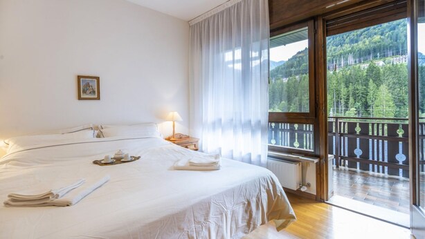 A hotel room with a large bed, a balcony with a view of a forested mountain, and a window with sheer curtains.