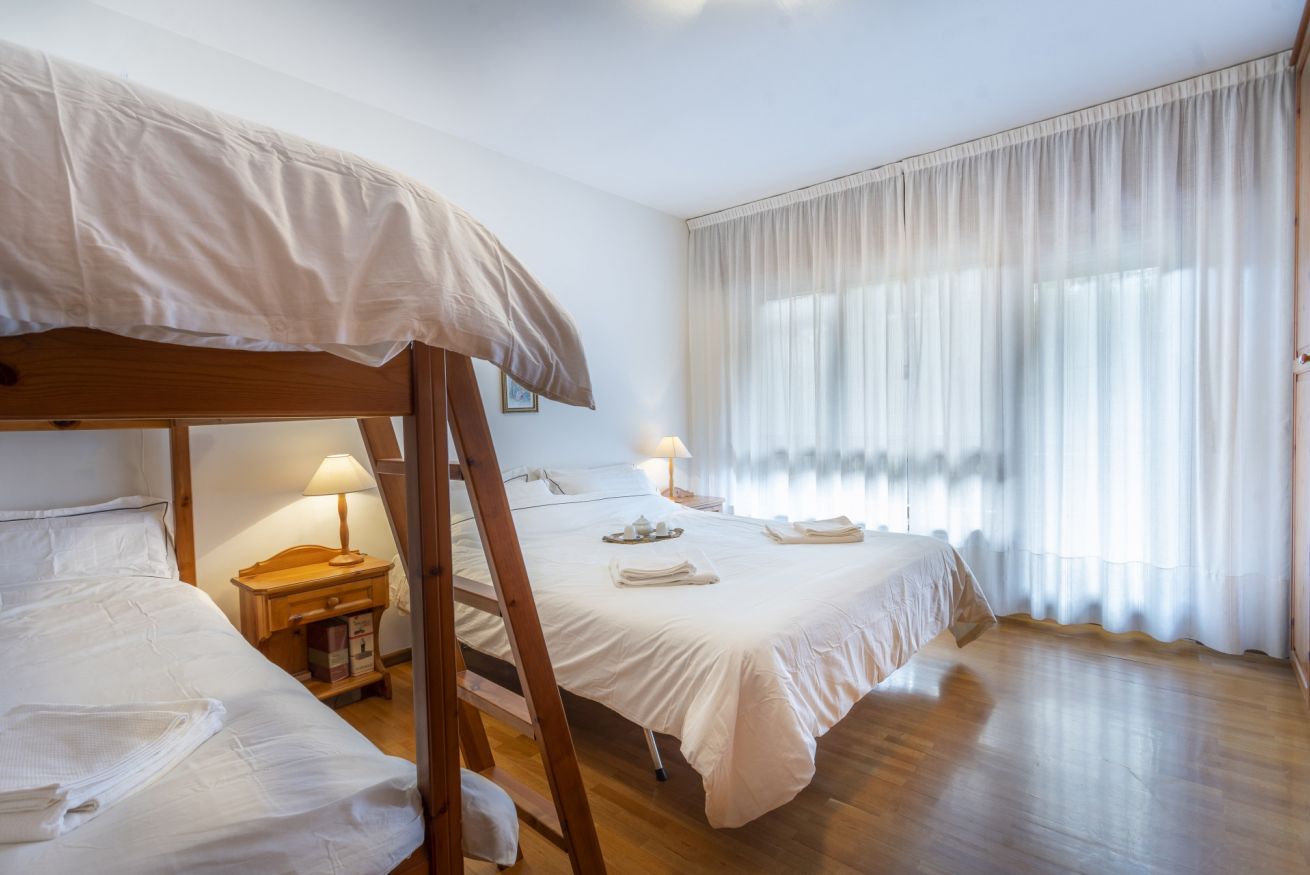 Ein Hotelzimmer mit Etagenbett, Nachttisch mit Lampe, Fenster mit durchsichtigen Vorhängen und Holzboden.
