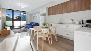 A modern kitchen with white cabinetry, a dining area with a wooden table and chairs, and a living area with a blue sofa and large windows. - Wentworthville 2 Bedrooms 2 Bathrooms with Free Parking by KozyGuru (Wentworthville)