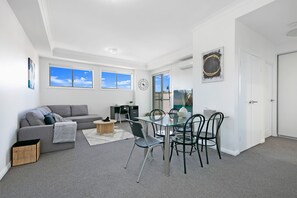 Ein modernes Wohnzimmer mit einem grauen Sofa, einem Glastisch mit Stühlen, einem kleinen Holznachttisch und einem großen Fenster mit Blick zum Himmel.