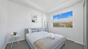 Ein modernes Schlafzimmer mit einem großen Bett, einem kleinen Nachttisch, einem Fenster mit Blick auf die Stadt und einer Deckenleuchte.