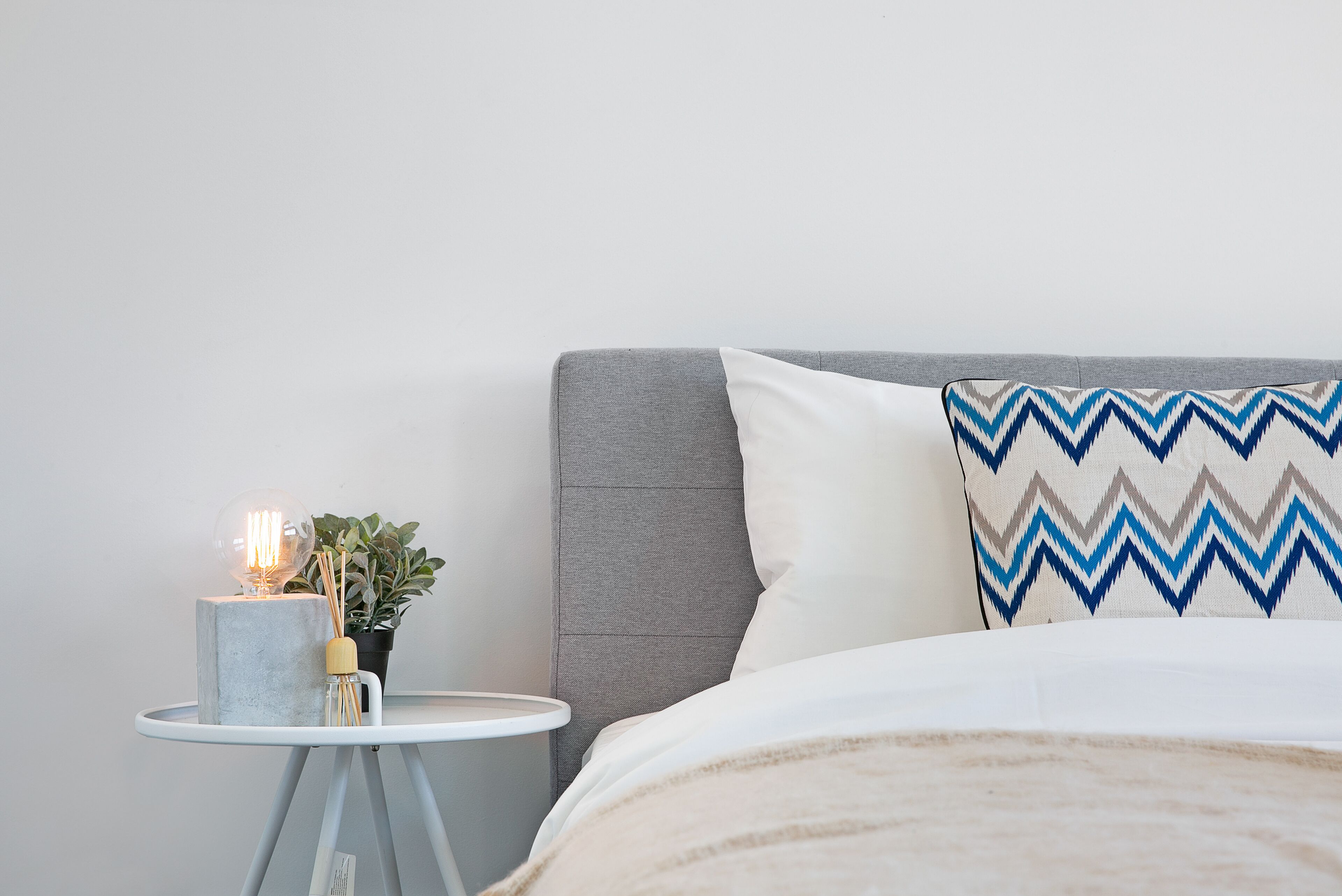 Ein ordentlich eingerichtetes Schlafzimmer mit grauem Kopfteil, einem weißen Kissen mit Zickzackmuster, einem kleinen runden Tisch mit Lampe und Topfpflanze sowie einer flauschigen, beige farbenen Decke auf dem Bett.