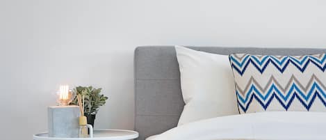 Une chambre à coucher bien rangée, avec une tête de lit grise, un oreiller blanc à motifs en zigzag, une petite table ronde avec une lampe et une plante en pot, et une couverture beige moelleuse sur le lit.