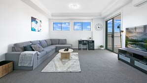 Ein modernes Wohnzimmer mit einem grauen Sofa, einem Couchtisch, einem Fernseher und einem großen Fenster mit Blick auf den Himmel.