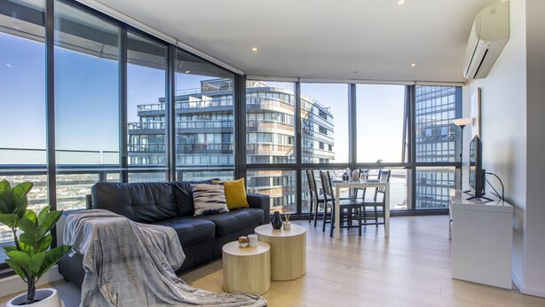 A modern living room with a large window, a sofa, a coffee table, and a dining area. - Docklands high level 1 Bedroom Apartment with pool by KozyGuru (Docklands)