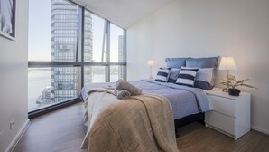 A modern bedroom with a large window, a bed with a blue headboard, a bedside table with a lamp, and a view of a cityscape. - Docklands high level 1 Bedroom Apartment with pool by KozyGuru (Docklands)