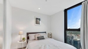 Une chambre à coucher moderne avec une grande fenêtre, un lit soigneusement fait, une petite table de chevet avec une lampe et un tableau encadré au mur.