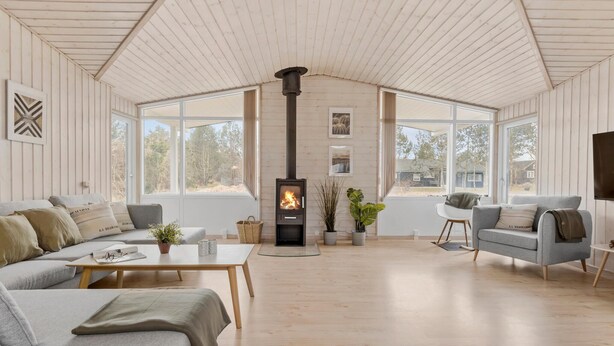 Un salón espacioso con techo alto, paredes de madera y ventanas amplias. Una estufa de leña está ubicada en el centro, y hay dos sofás y una mesita.