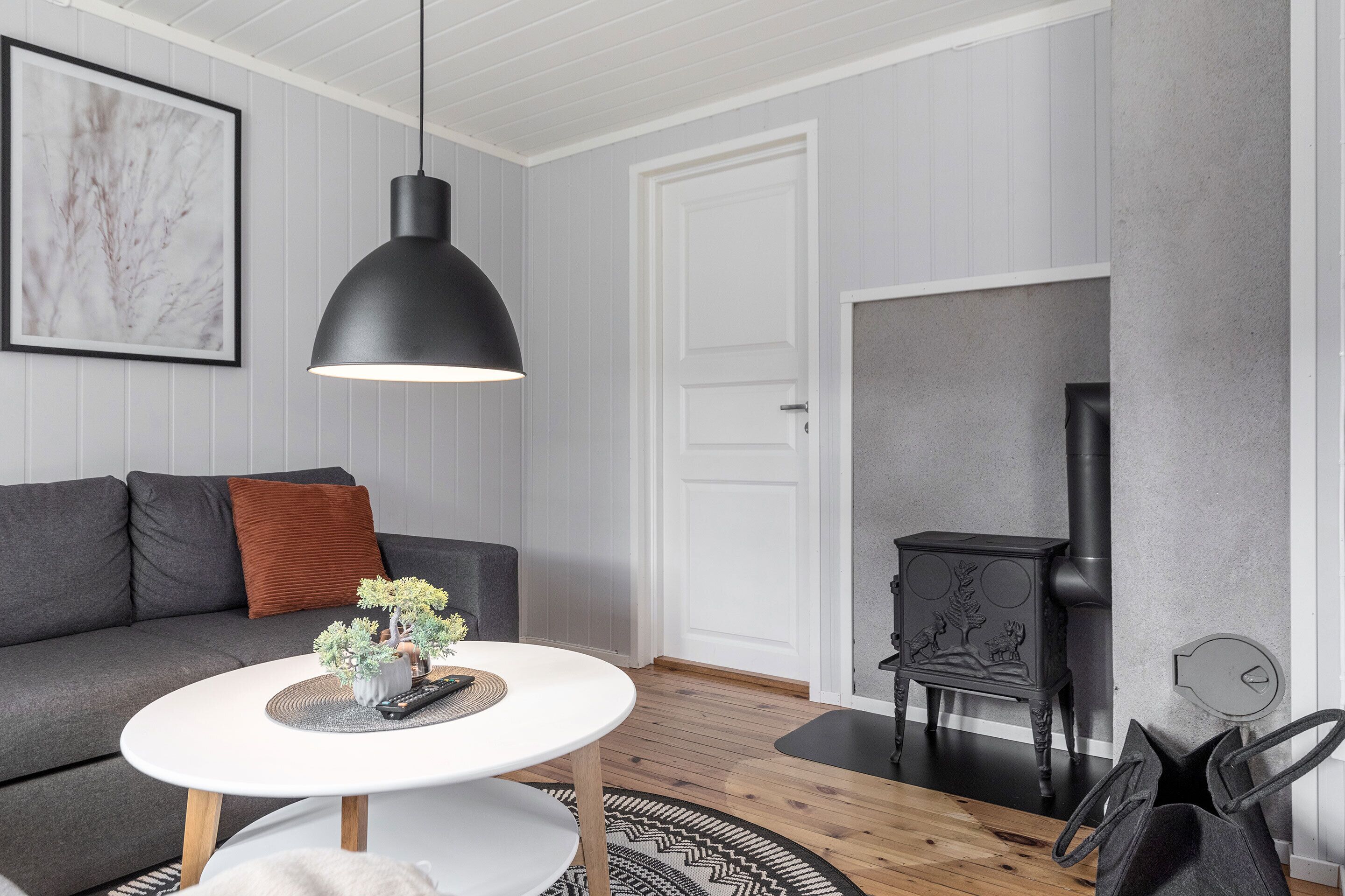 A modern living room with a grey sofa, a white coffee table, a black pendant light, a black stove, and a white door.