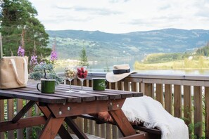 Una terraza de madera con una mesa de picnic, una taza verde, un vaso con bayas, un sombrero de paja y un bolso tejido.