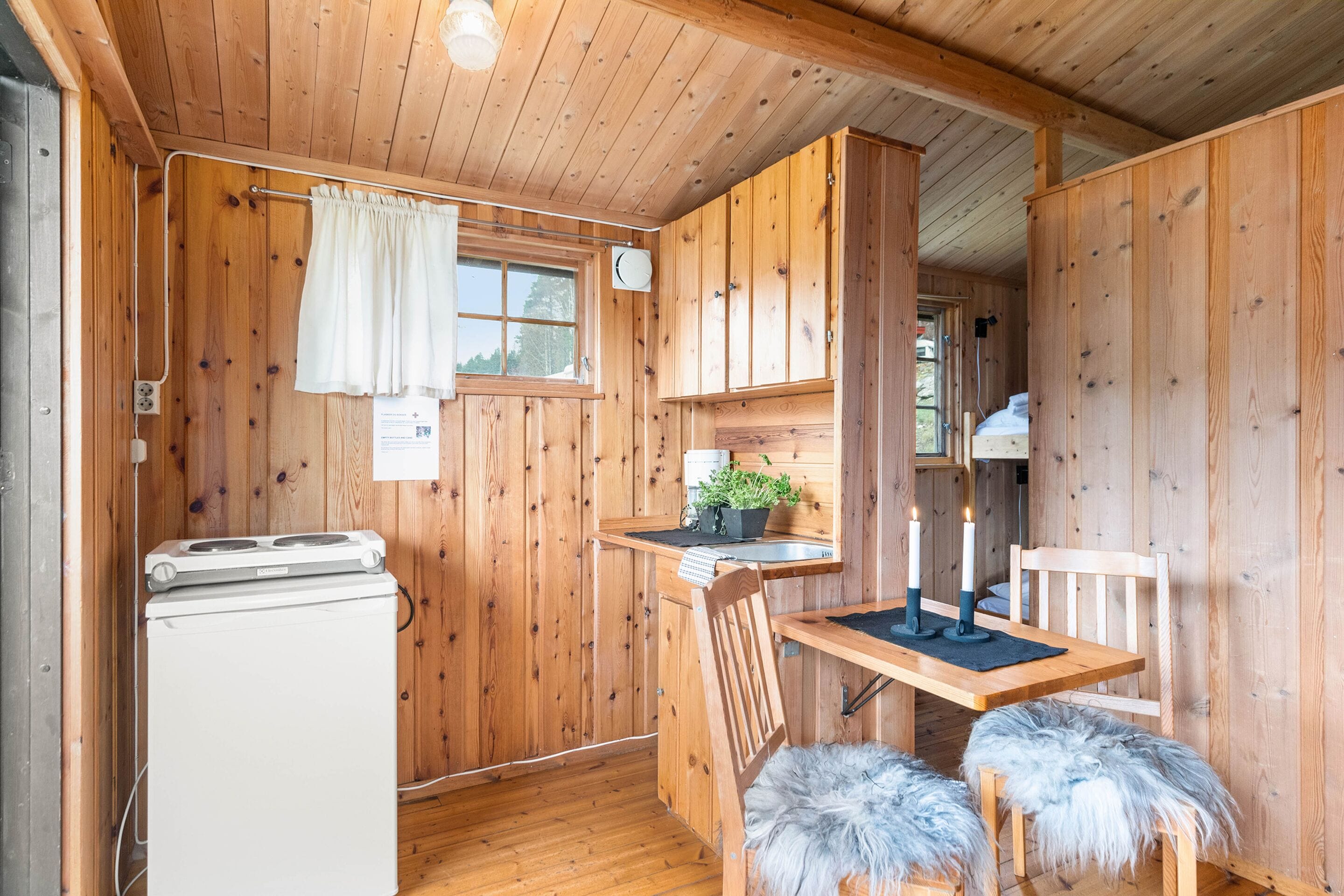 A wooden cabin interior with a kitchen area, including a refrigerator, a dining table with two chairs, and a window with curtains.