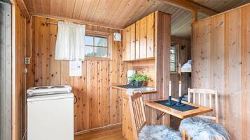 A wooden cabin interior with a kitchen area, including a refrigerator, a dining table with two chairs, and a window with curtains.