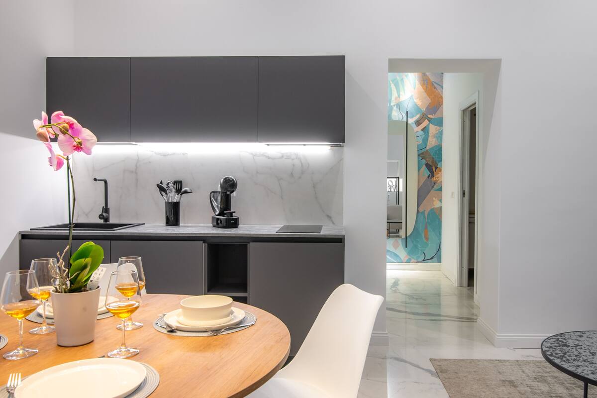A modern kitchen with dark cabinets, a marble countertop, and a dining area with a wooden table and white chairs.