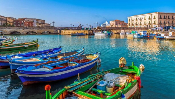 Un port où sont amarrés plusieurs bateaux aux couleurs vives, un pont de pierre et des bâtiments en arrière-plan.