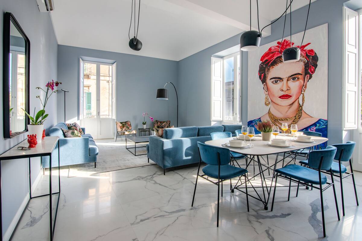 A modern living room with a blue sofa, a dining table with chairs, and a large window.